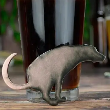 Novelty dog-shaped bottle opener leaning on a glass of dark beer on a wooden table.