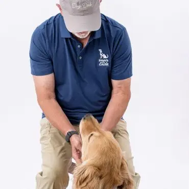 Volunteer with a golden retriever, wearing a Dog Dad cap and Paws With A Cause shirt, interacting lovingly.