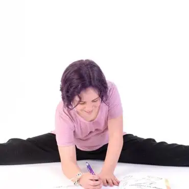 Child writing in a book while doing a split on the floor, wearing a pink shirt and black pants.