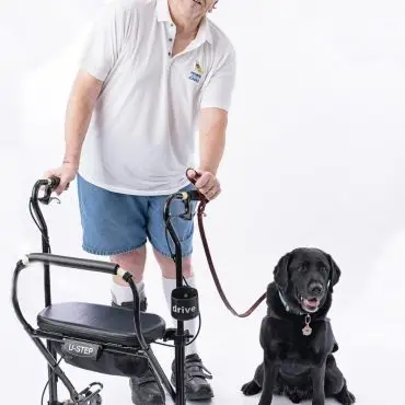 Man with a walker and a black service dog on a leash, smiling against a white background.