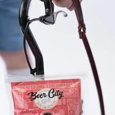 Hand holding a dark bicycle handle with Beer City dog biscuits attached, showcasing pet treats on the go.