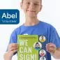 Boy in blue shirt holding We Can Sign! book on sign language for kids, smiling. Text: Abel Volunteer.