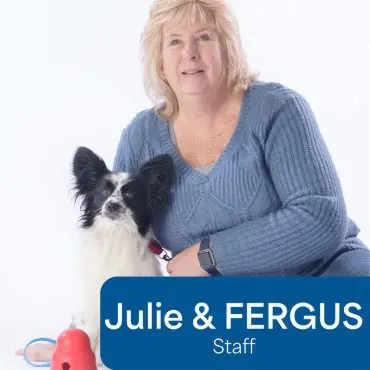 Blonde woman in a blue sweater poses with a black and white dog named Fergus, next to a red dog toy, on a white background.