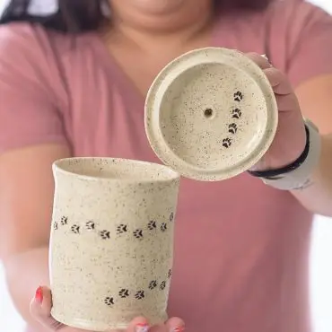 Person holding speckled pottery cup and lid with paw print design, showcasing handmade ceramic craftsmanship.