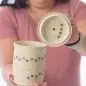 Person holding speckled pottery cup and lid with paw print design, showcasing handmade ceramic craftsmanship.