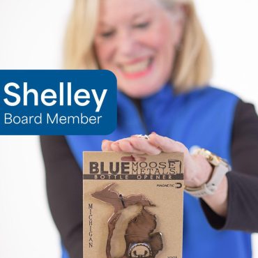 Board member Shelley displays a Michigan-shaped metal bottle opener by Blue Moose Metals, smiling in a blue vest.