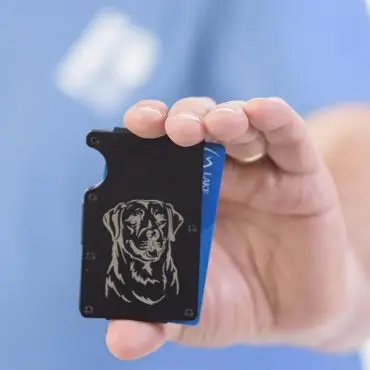 Hand holding black wallet with engraved dog design on front, blue background.