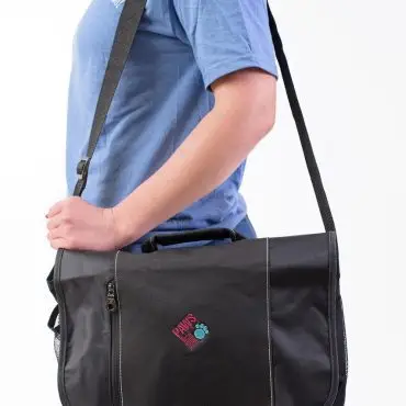 Woman wearing a blue shirt carrying a black messenger bag with logo, over white background.