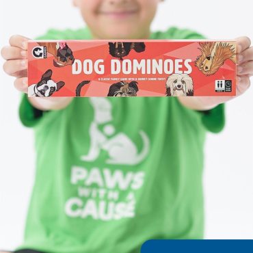 Child in green shirt holds Dog Dominoes game box, promoting a family-friendly canine-themed board game.