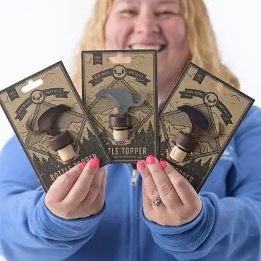 Smiling person holding three Blue Moose Metals bottle toppers, wearing a blue hoodie.