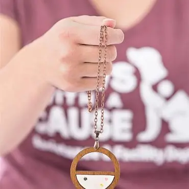 Hand holding a wooden round pendant necklace with a minimalist design, worn with a purple shirt.