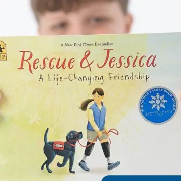 Boy holding 'Rescue & Jessica' book about girl and service dog, featuring award sticker and Teddy Volunteer badge.