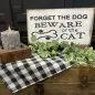 Decorative sign saying Forget the Dog, Beware of the Cat with candle and greenery on a checkered tablecloth.