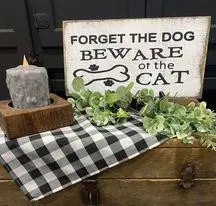 Decorative sign saying Forget the Dog, Beware of the Cat with candle and greenery on a checkered tablecloth.