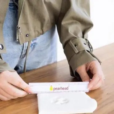 Person measuring heart-shaped clay with ruler on wooden table, wearing green jacket and jeans.