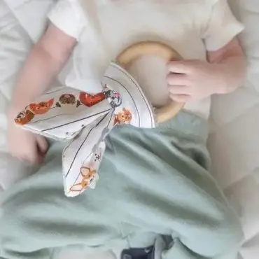 Baby holding a wooden teething ring with fabric ears, wearing a white shirt and green pants on a quilted surface.