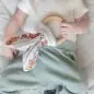 Baby holding a wooden teething ring with fabric ears, wearing a white shirt and green pants on a quilted surface.