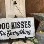 Decorative sign saying Dog Kisses Fix Everything with rustic decor, candle, and flowers on a wooden background.