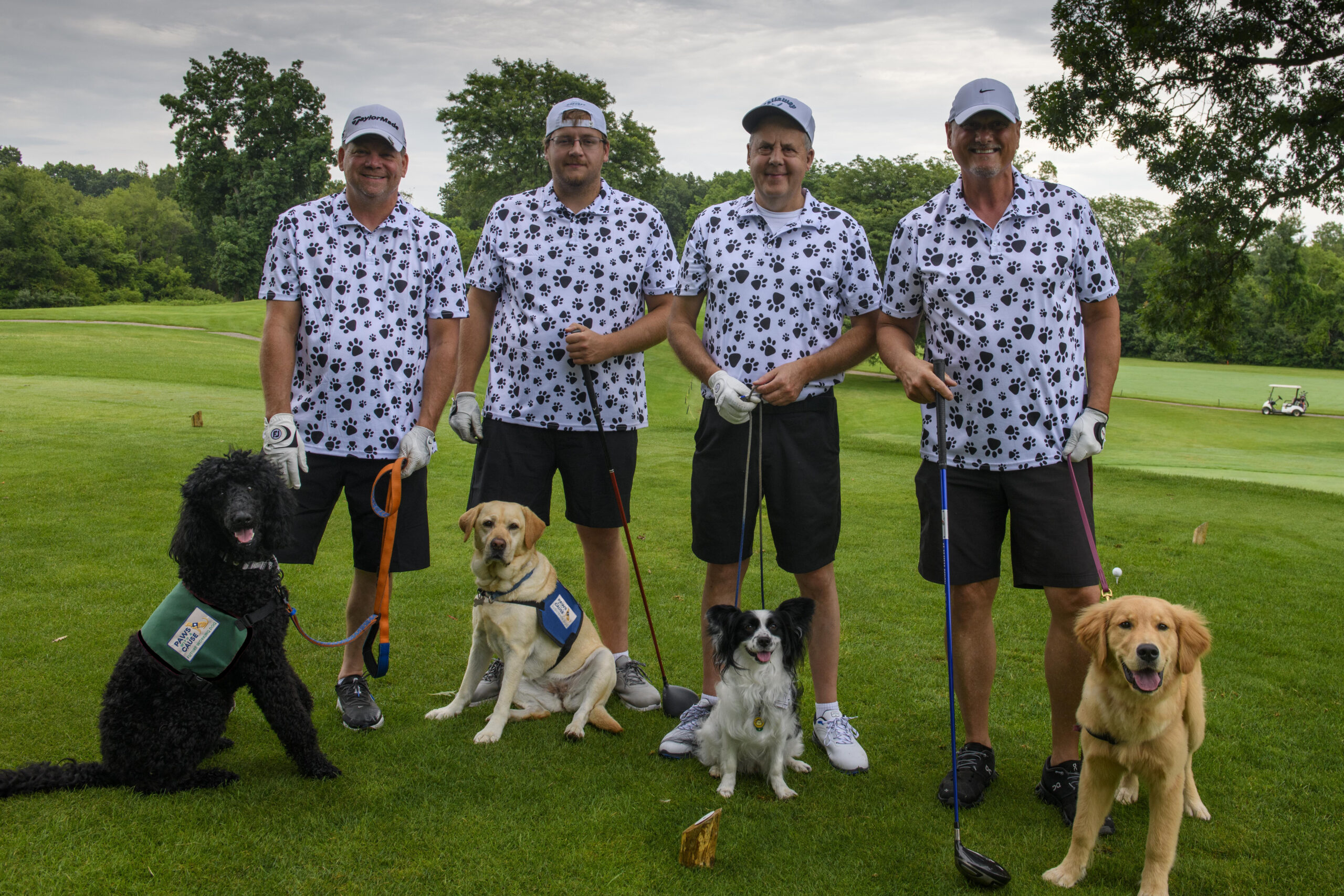 Four golfers with matching paw-print shirts and three dogs on a golf course.