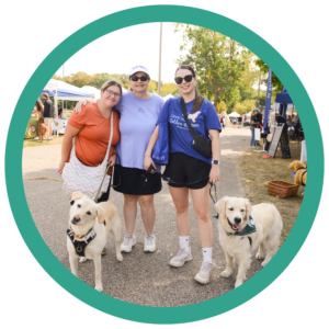 Three women with two golden retrievers at an outdoor event on a sunny day.