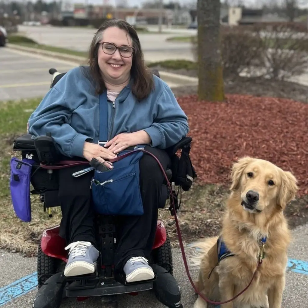 Smiling person in wheelchair with service dog on a path, surrounded by greenery and mulch.