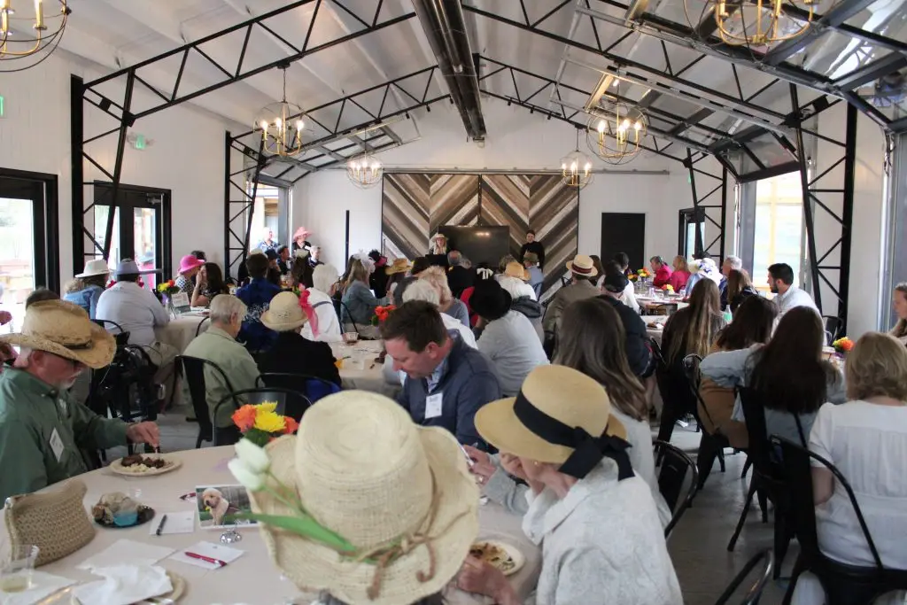 People wearing hats enjoying a social gathering with dining tables and chandeliers in a modern rustic setting.