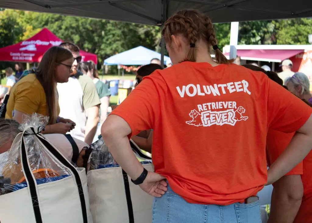 Volunteer event with people in orange shirts, exchanging gift bags outdoors under tents on a sunny day.