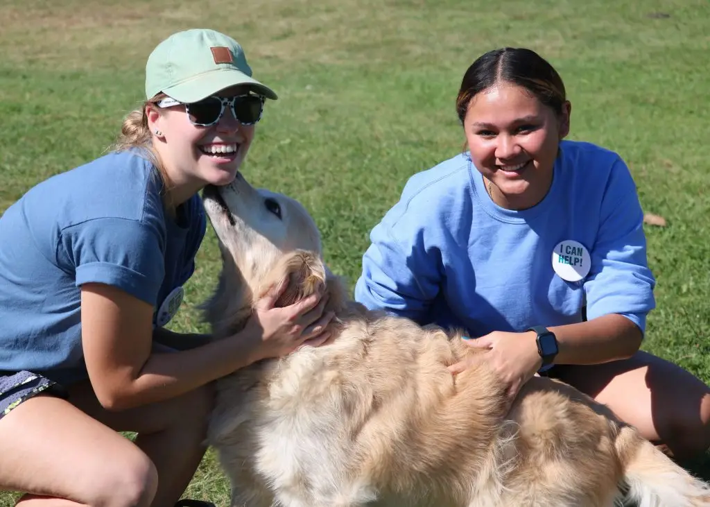 Two people smiling with a golden retriever outdoors on a sunny day.
