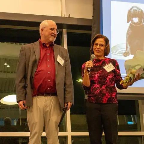 Man and woman speaking at an event, holding a microphone and bouquet, with a dog image projected in the background.