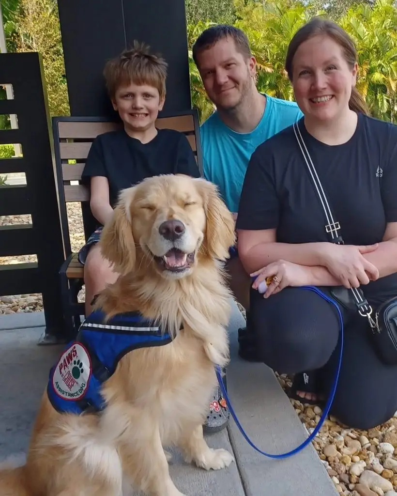 Family with a smiling service dog in blue vest sitting outdoors.