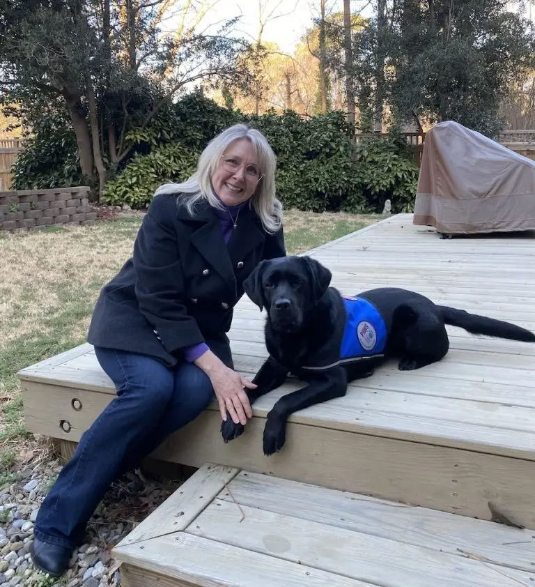 Woman sitting on a deck with a black service dog, surrounded by greenery in a backyard setting.