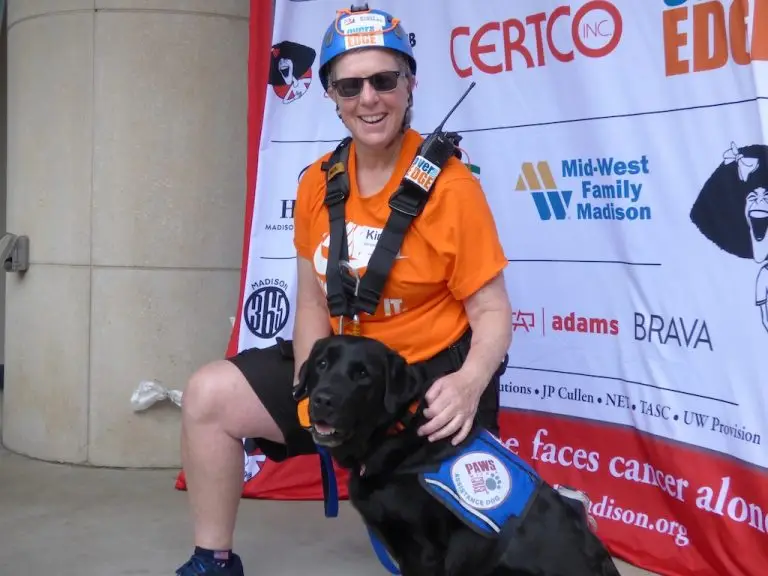 Person in climbing gear smiling with a service dog at a charity event backdrop.