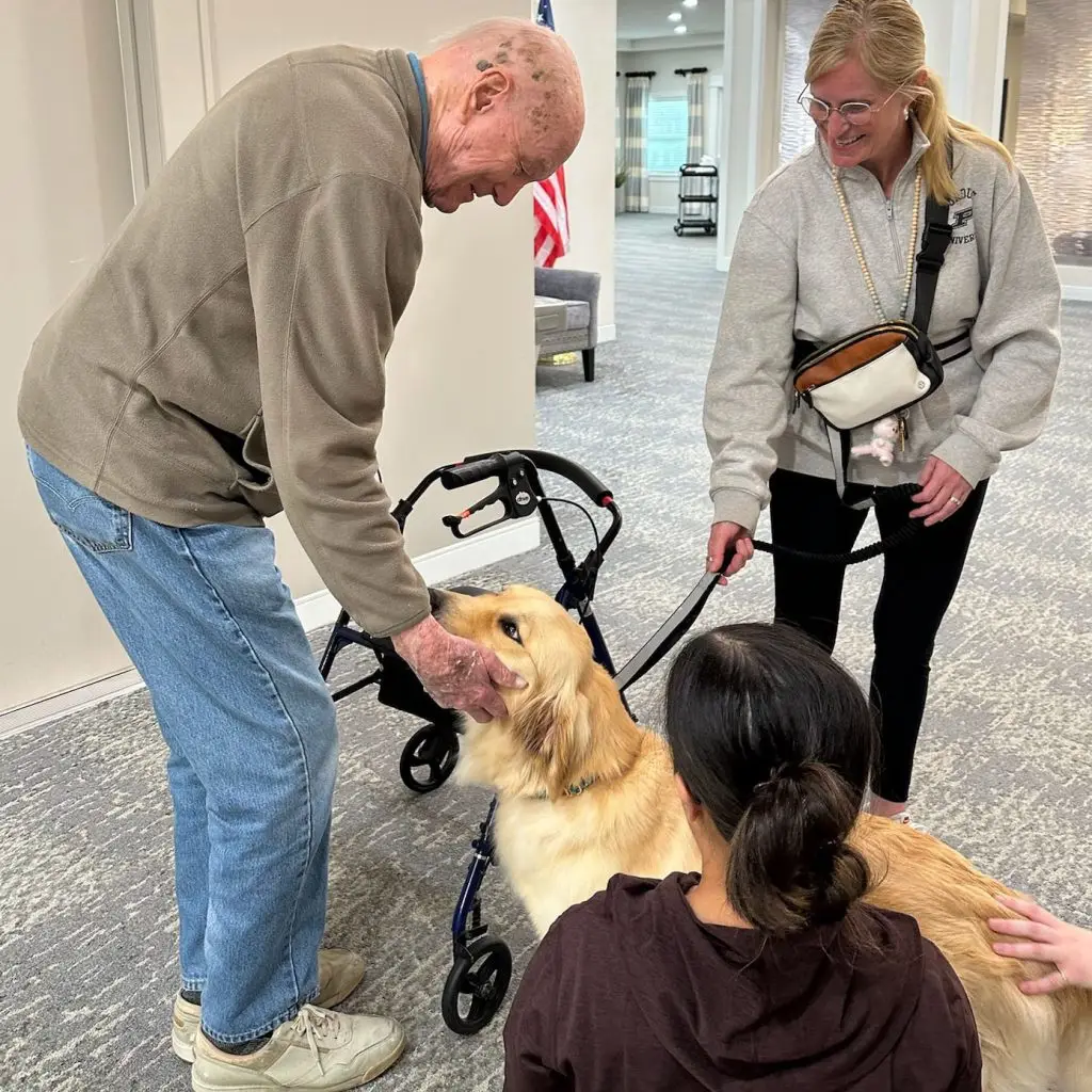 Elderly man petting a golden retriever with a walker, accompanied by two women in a care facility.