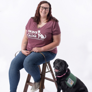 Volunteer with therapy dog sitting on wooden stool, wearing Paws with a Cause shirt.