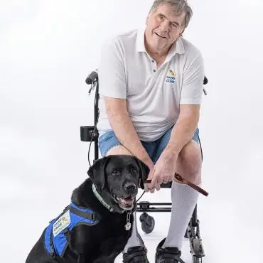 Man in wheelchair with assistance dog in blue vest, both smiling. Empowerment and support through service animals.