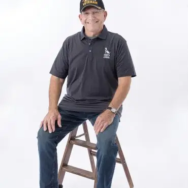 Smiling veteran wearing a dark cap and shirt sits on a wooden stool against a white background.