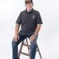 Smiling veteran wearing a dark cap and shirt sits on a wooden stool against a white background.