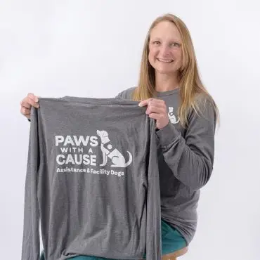Smiling woman holds a Paws with a Cause shirt, promoting assistance and facility dogs.
