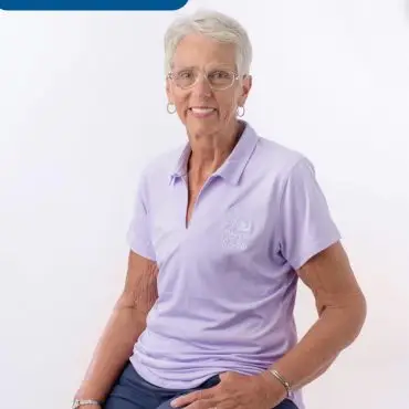 Volunteer named Connie sitting on a stool, wearing a lavender Paws for Cause polo shirt, smiling at the camera.