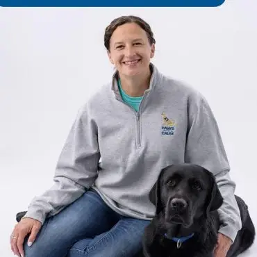 Smiling woman in grey sweater with black Lab on white background, Paws for a Cause logo visible.