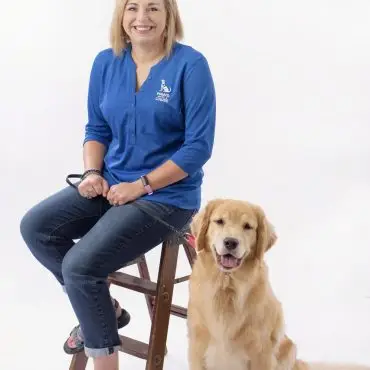 Smiling woman in blue shirt with golden retriever sitting on a stool against a white background.
