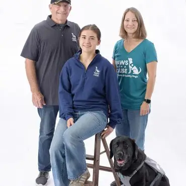 Group with a service dog wearing Paws With A Cause shirts, posing together. White background.
