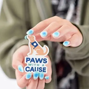 Person holding Paws with a Cause sticker with blue-tipped nails, promoting animal charity support.