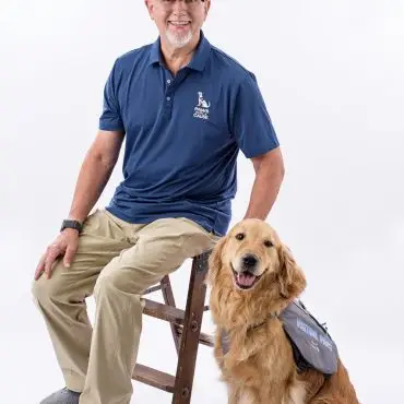 Man sitting with a therapy dog wearing a Visiting Paws vest, smiling together in a studio setting.