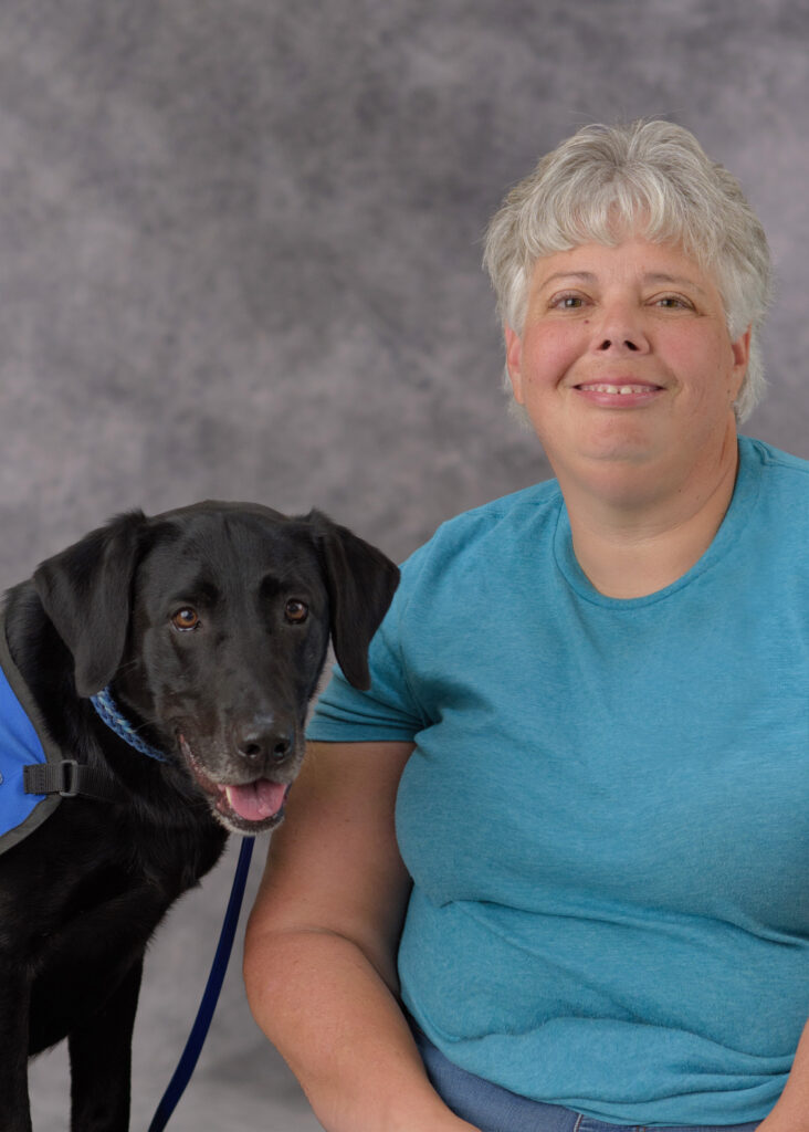 Person smiling beside a black Labrador wearing a blue vest, studio portrait with gray background.