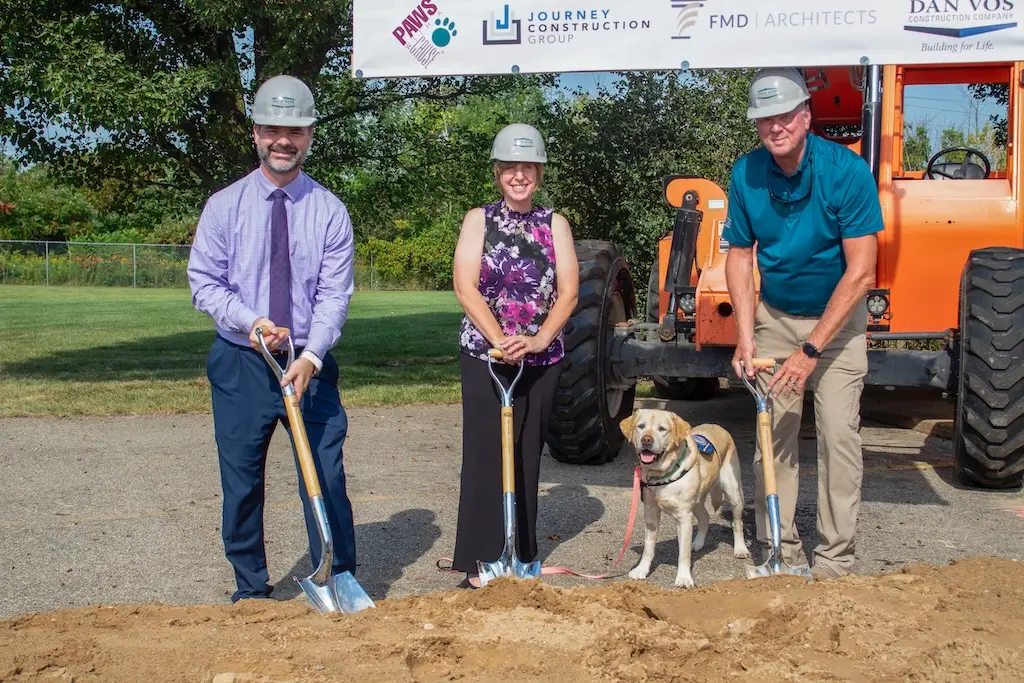 People and a service dog participate in a groundbreaking ceremony with shovels, construction equipment in the background.