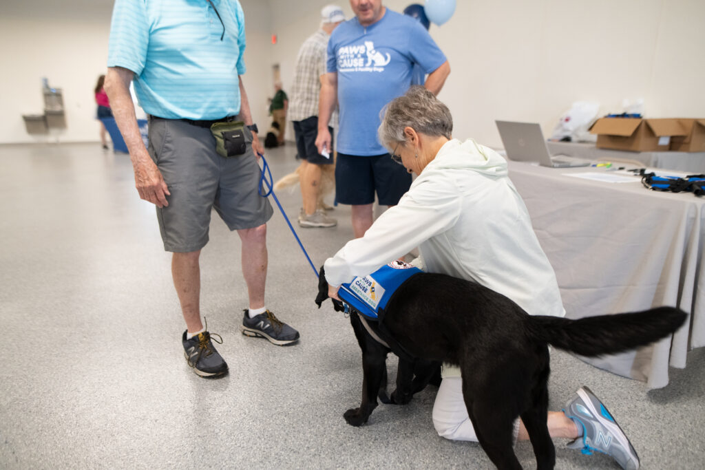 Elderly woman meeting a black service dog at an event, surrounded by people in casual attire.
