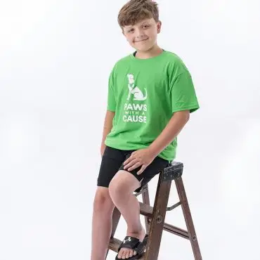 Boy in green Paws with a Cause shirt sits on a ladder against white background, promoting animal charity.