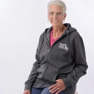 Elderly woman smiling, wearing a grey hoodie with Paws Cause logo, seated on a wooden stool against a white background.