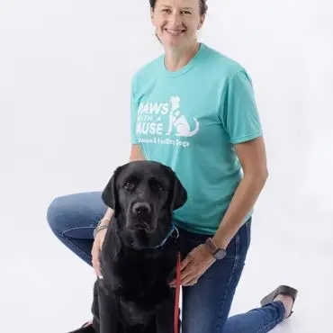Woman kneeling with a black Labrador wearing a service dog tee, on white background.